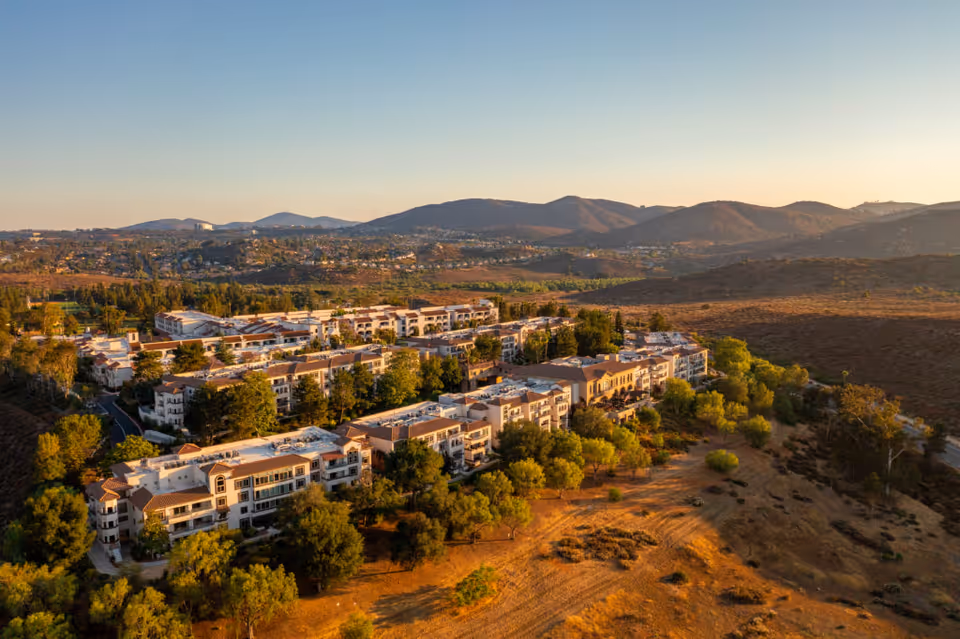 Aerial view of Casa de las Campanas senior living facility surrounded by trees and dry hills, with mountains in the background under a clear sky during sunset.
