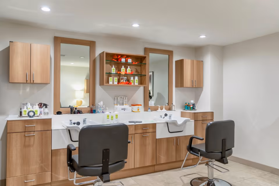 A clean and modern hair salon area with two black salon chairs facing two white sinks. The wall behind has wooden cabinets, two large mirrors, and shelves stocked with hair care products and decorations. The room is well-lit with recessed ceiling lights and has a tiled floor.