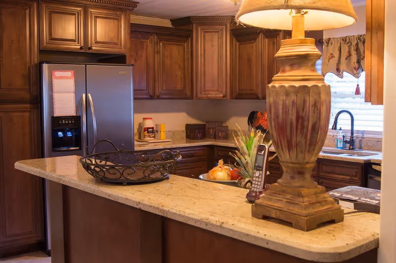 A warm, well-lit kitchen with wooden cabinets, a stainless steel refrigerator, a granite countertop island with a decorative basket, a lamp, and various kitchen items including a pineapple and an onion. A window with blinds and a floral valance is above the sink.
