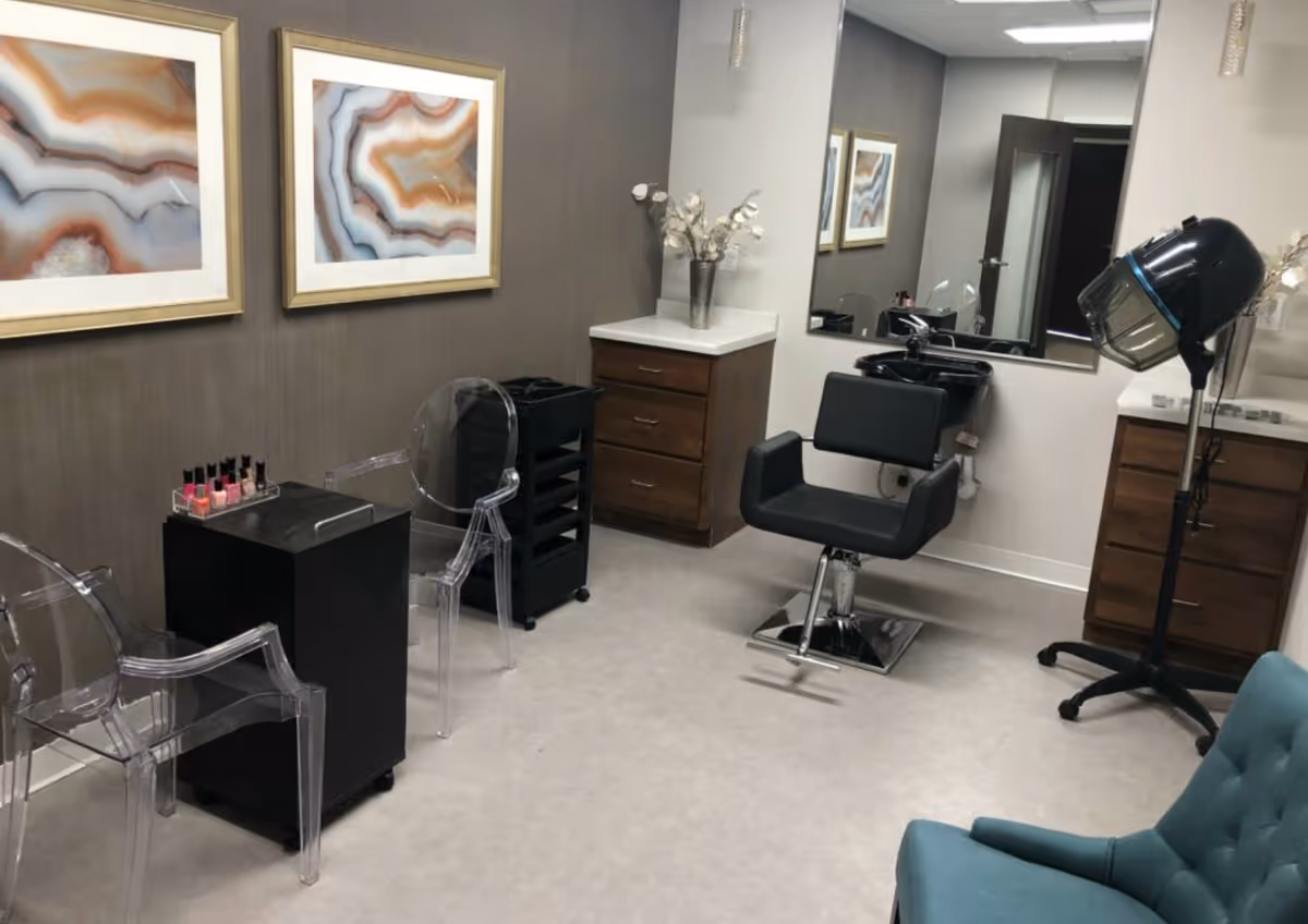 A small salon room with a black salon chair in front of a mirror and a black hair washing sink. There is a black hair dryer on a stand, two clear acrylic chairs, a small black table with nail polish bottles, and two framed abstract art pieces on the wall. The room has light-colored flooring and neutral-toned walls with wooden cabinets and a blue cushioned chair in the corner.