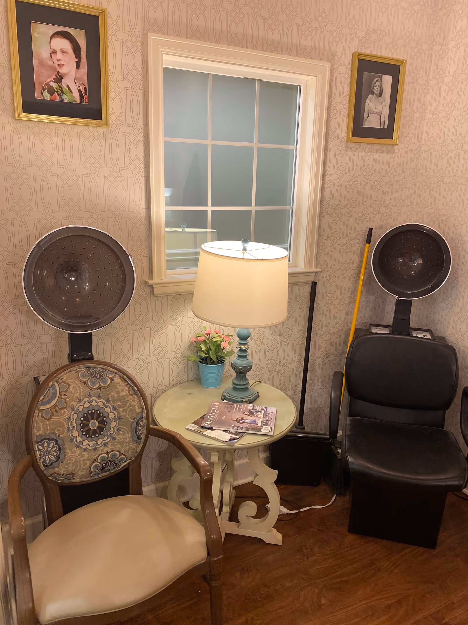 A cozy salon corner with two vintage hair dryer chairs, one with a patterned cushion and the other black leather. Between them is a small round table with a blue lamp, a small potted plant, and magazines. The wall behind has patterned wallpaper and two framed black-and-white portraits. A window with white trim is centered on the wall.