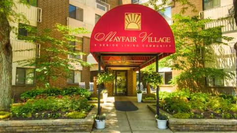 Front entrance of Mayfair Village Retirement Center with red canopy and landscaped walkway.