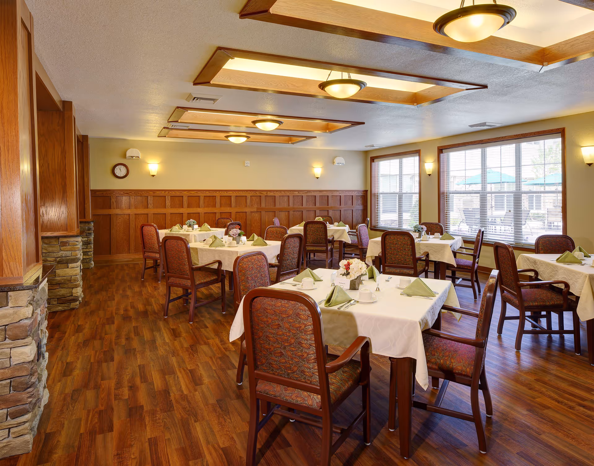 Well-lit dining room with multiple tables set with white tablecloths, green napkins, and chairs in a senior living facility.
