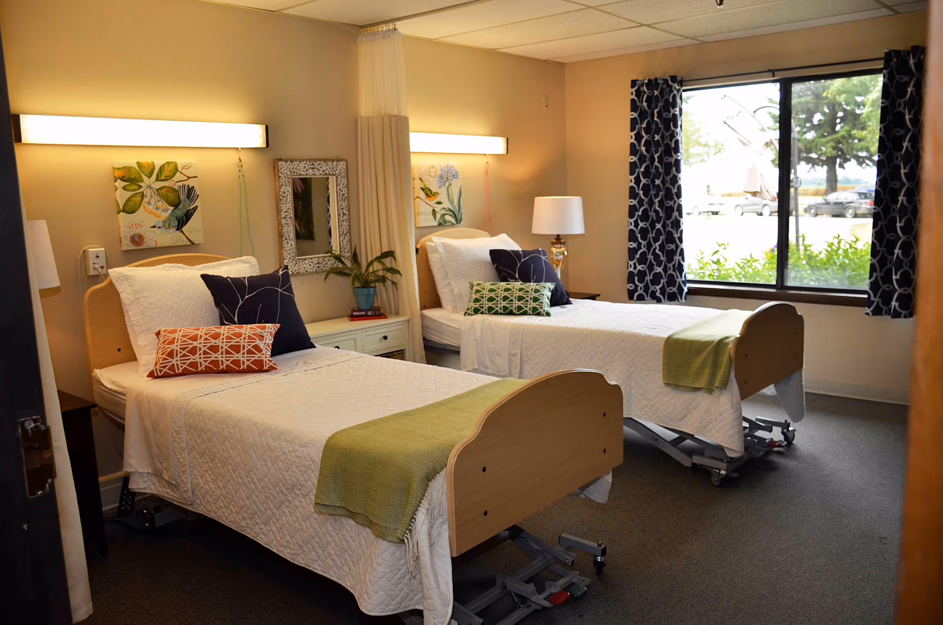 A bright room with two single hospital-style beds, each with white bedding, colorful pillows, and a green throw blanket. Between the beds is a small white nightstand with a potted plant and a lamp. The room has a large window with patterned curtains, letting in natural light and showing some greenery and parked cars outside. The walls are decorated with framed artwork and a mirror.