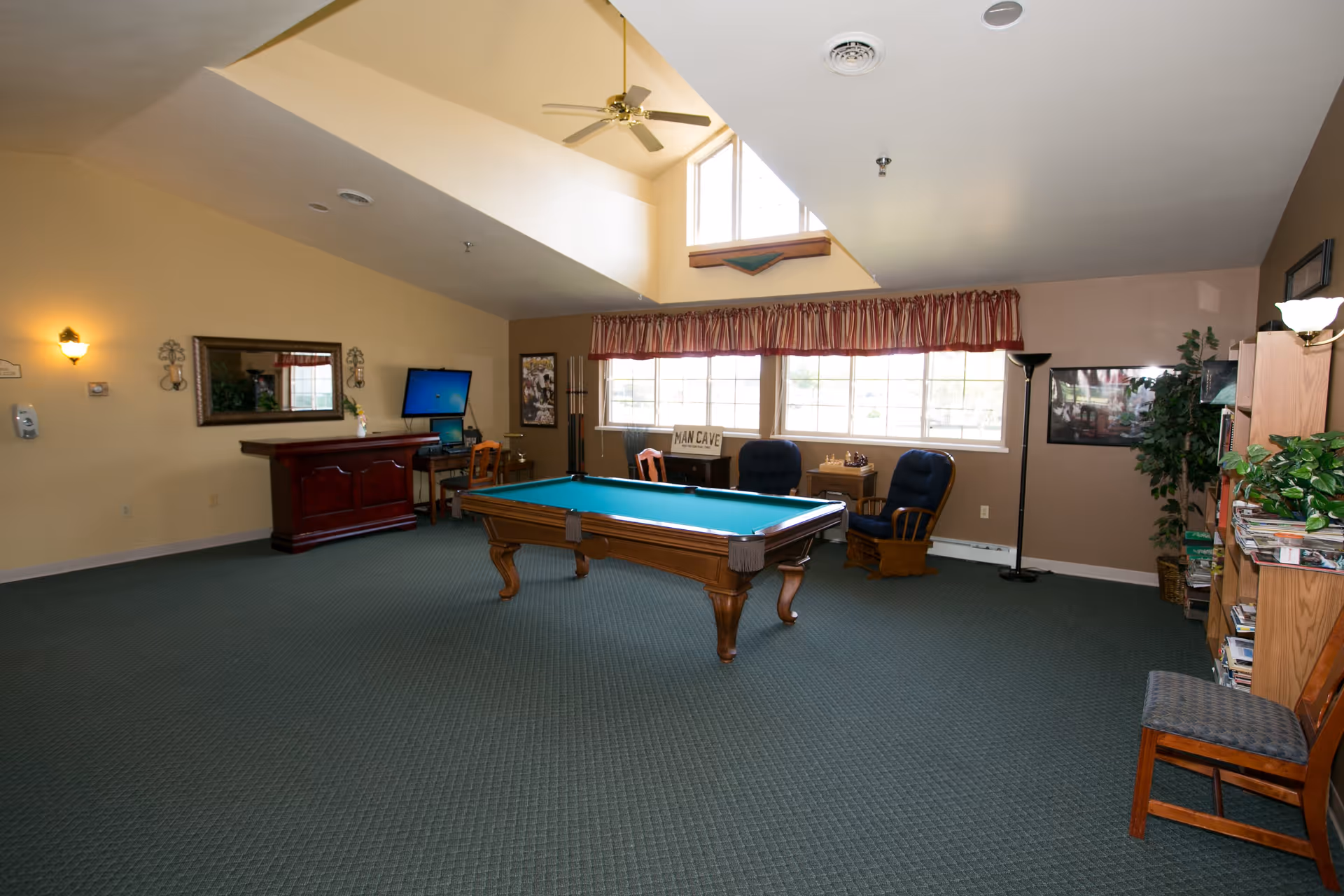 A spacious recreational room with a pool table in the center, two cushioned rocking chairs near a window with red and white striped valance, a small table with a chess set, a computer desk with a monitor, a wooden bar, bookshelves filled with books and plants, and a ceiling fan above. The room has carpeted flooring and beige walls.