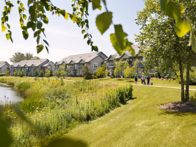 A scenic outdoor view of ProHealth Regency Senior Communities Muskego featuring a grassy area with trees and shrubs, a walking path with three people strolling, and a row of residential buildings in the background under a clear sky.