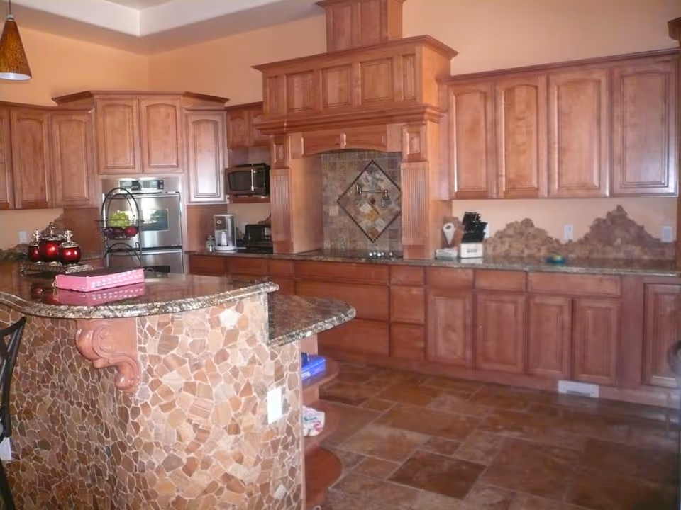 Spacious kitchen with wooden cabinets, a tiled curved island and granite countertops with appliances.