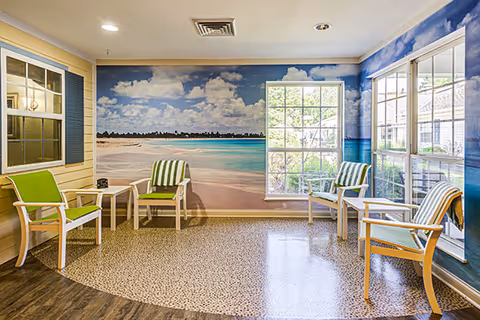A bright room with a tropical beach mural covering two walls, featuring blue skies, clouds, and ocean waves. The room has four white chairs with green and white striped cushions arranged around two small white tables. The floor has a mix of wood and pebble-like patterns. Large windows let in natural light and show greenery outside.