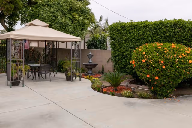 Outdoor patio area with a beige canopy covering a table and chairs, surrounded by potted plants and greenery. There is a multi-tiered water fountain against a brick wall, and a well-maintained garden with flowering bushes and shrubs.