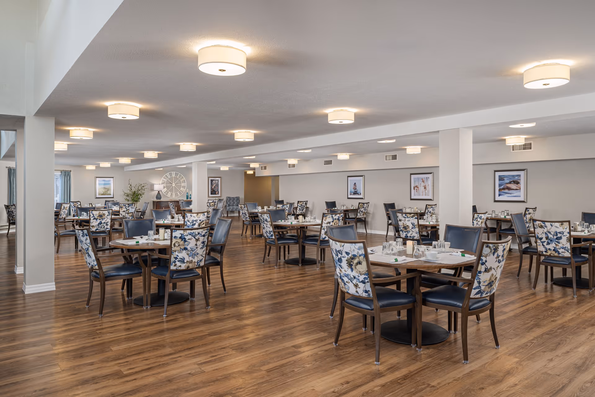 A spacious senior living dining room with multiple round tables, each surrounded by chairs with floral patterned backs and dark seats. The room has wooden flooring, soft overhead lighting, and framed artwork on the walls. The tables are set with glasses, napkins, and utensils, ready for a meal service.
