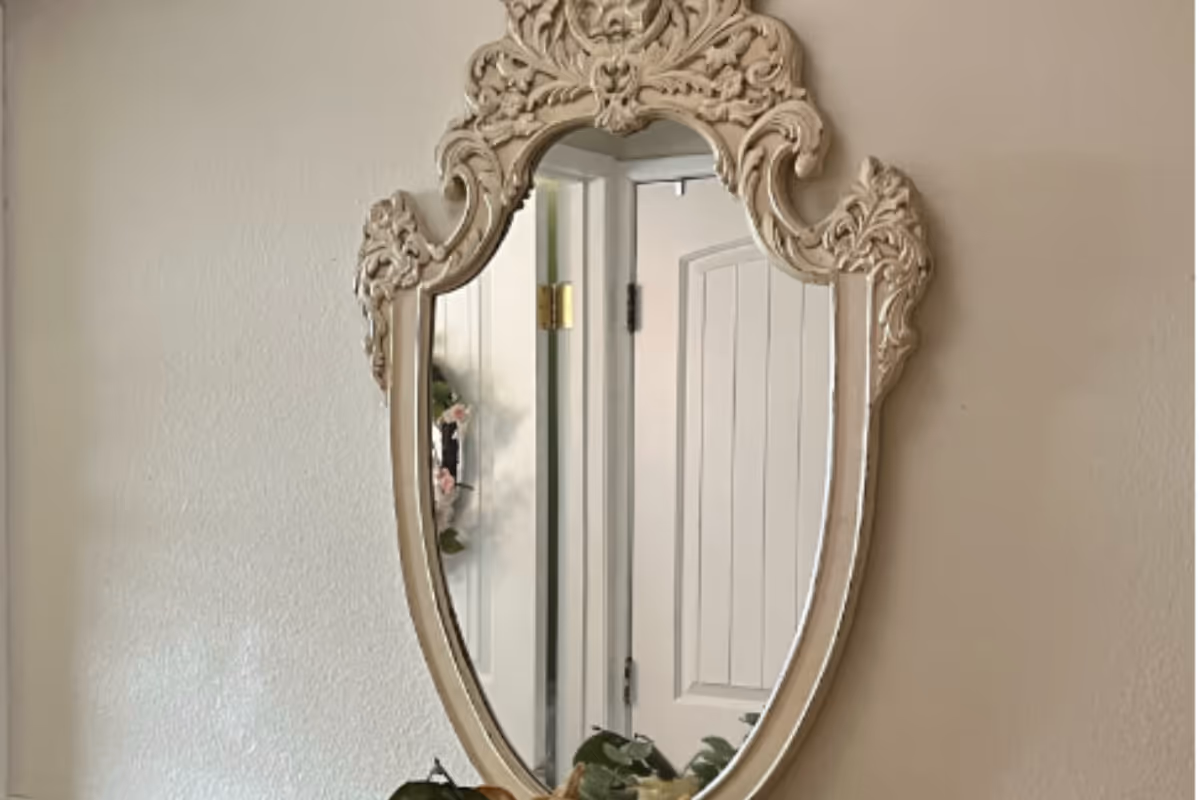 Ornately carved wall mirror reflecting a doorway and a small wreath, with a plant partially visible at the bottom.