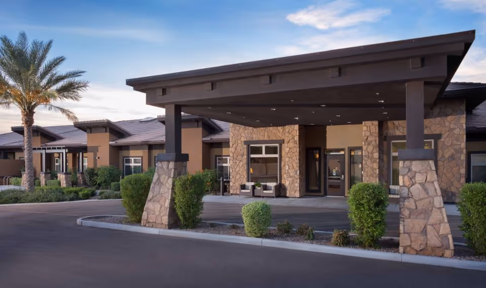 Exterior view of Mosaic Gardens Memory Care at Chandler showing the entrance with a covered driveway supported by stone pillars, surrounded by trimmed bushes and palm trees under a partly cloudy sky.
