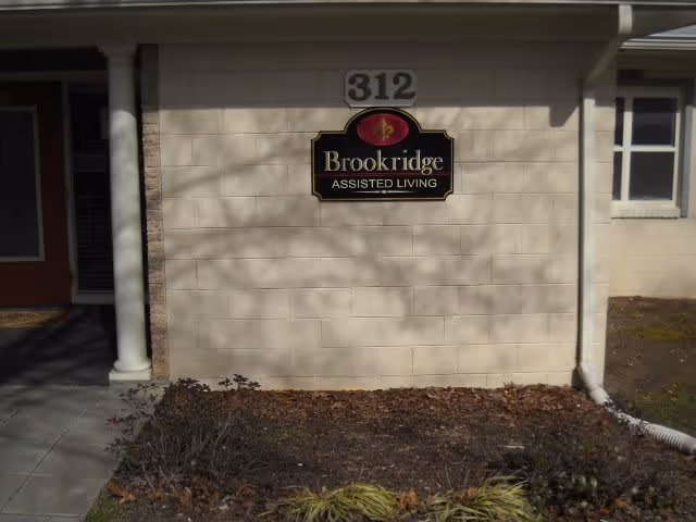 Exterior wall of a building with a sign that reads 'Brookridge Assisted Living' mounted below the number 312. The wall is light-colored with a small garden bed in front containing some plants and mulch. There is a white column on the left side and a window on the right side.
