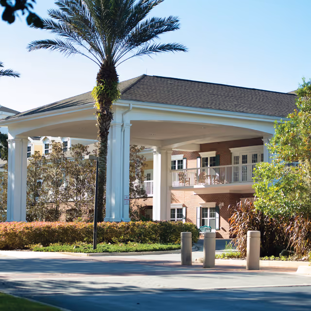 Exterior view of a senior living facility with a covered entrance supported by white columns, palm trees, and landscaped bushes. The building has brick walls and balconies with white railings.