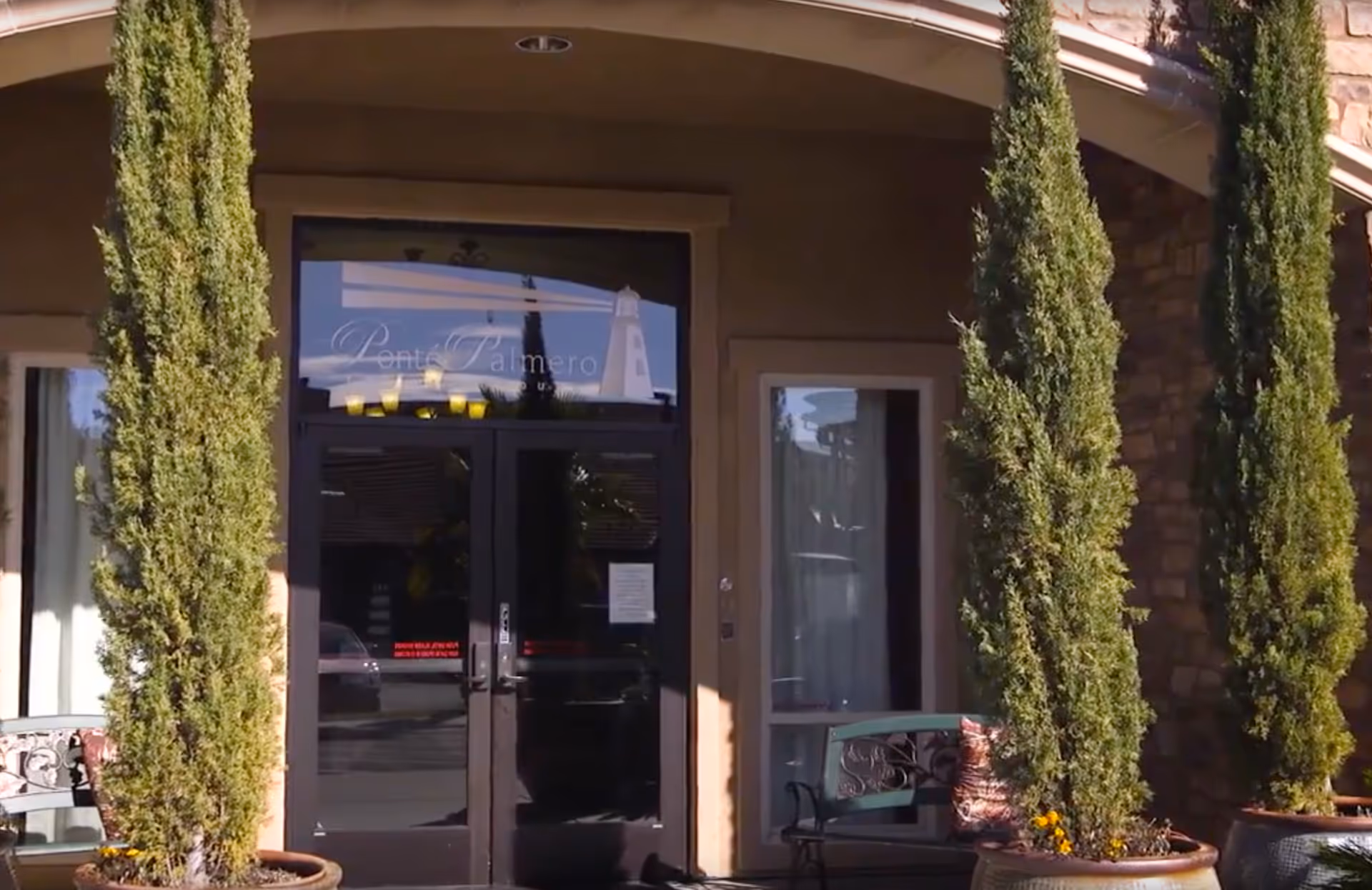 Entrance to Ponté Palmero facility with double glass doors, flanked by tall potted evergreen trees and a bench on the right side.