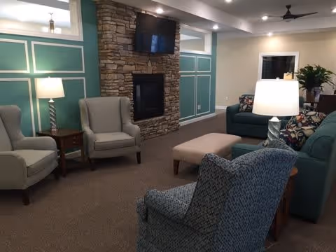 A cozy living room with a stone fireplace and a mounted flat-screen TV above it. The room features a mix of upholstered armchairs and sofas arranged around a rectangular ottoman. There are table lamps on side tables providing warm lighting, and the walls have decorative paneling painted in teal and beige tones.