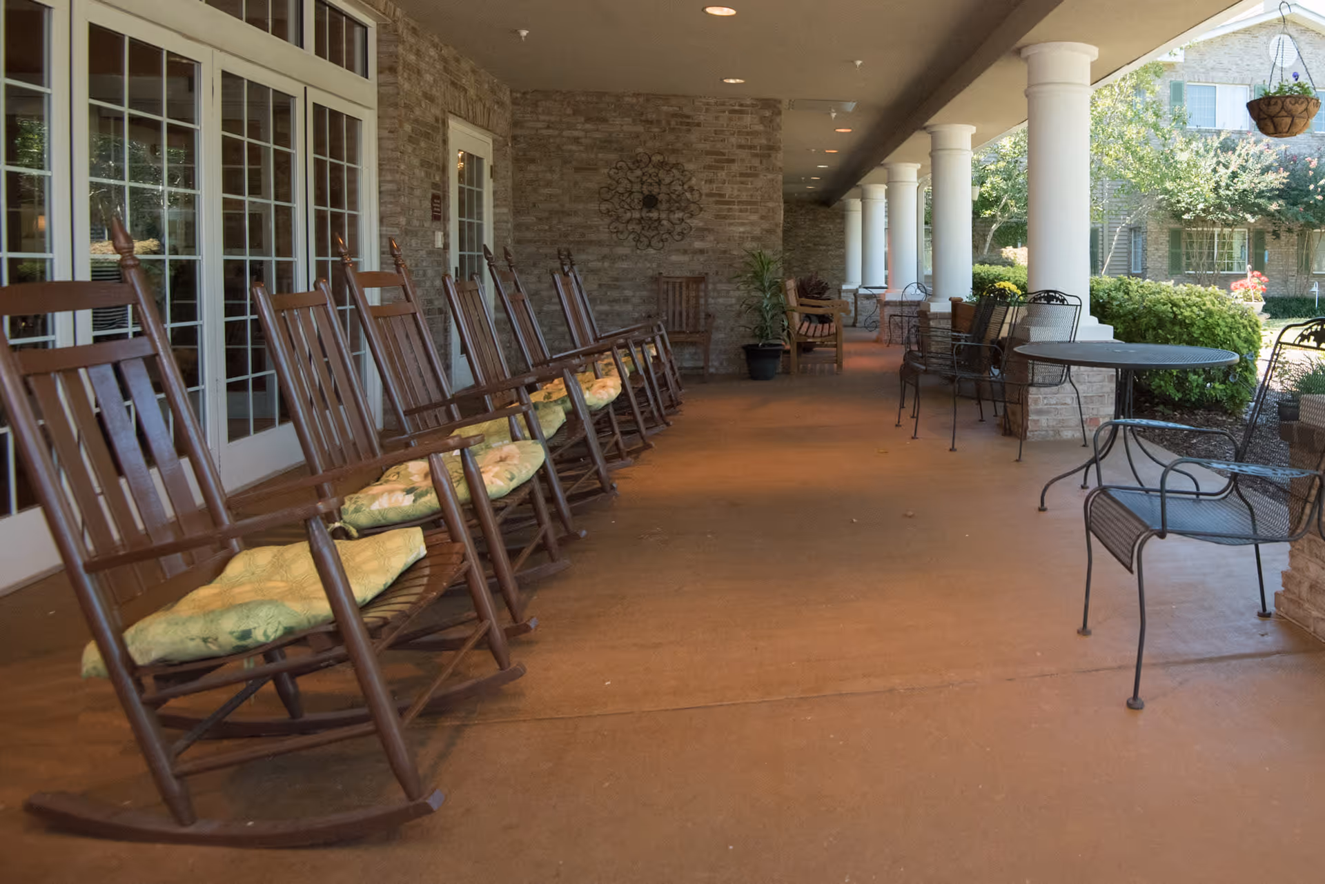 Covered outdoor patio area with a row of wooden rocking chairs with cushions on the left side, metal tables and chairs on the right side, white columns supporting the roof, and greenery visible beyond the patio.