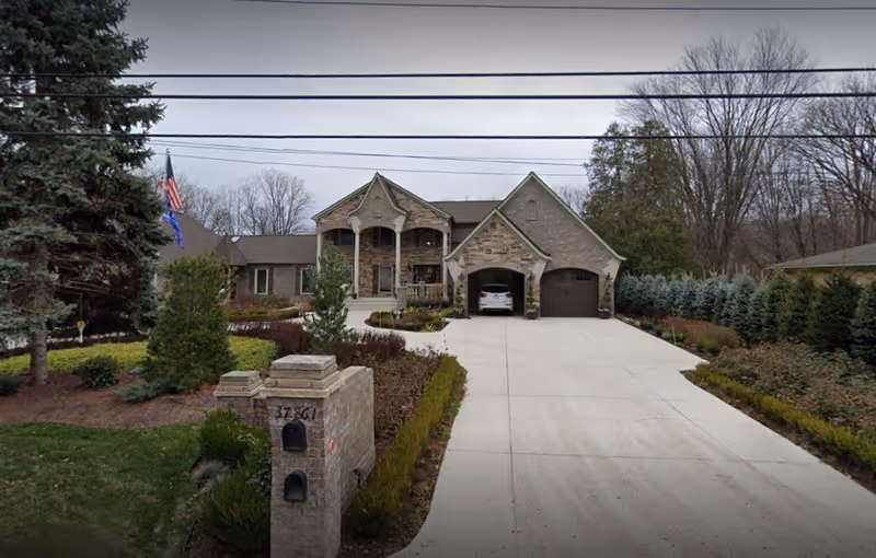 Front exterior view of a large brick house with a wide concrete driveway leading to a two-car garage. The house has a covered porch with columns and is surrounded by landscaped gardens and trees. An American flag and another flag are visible on the left side near the house.