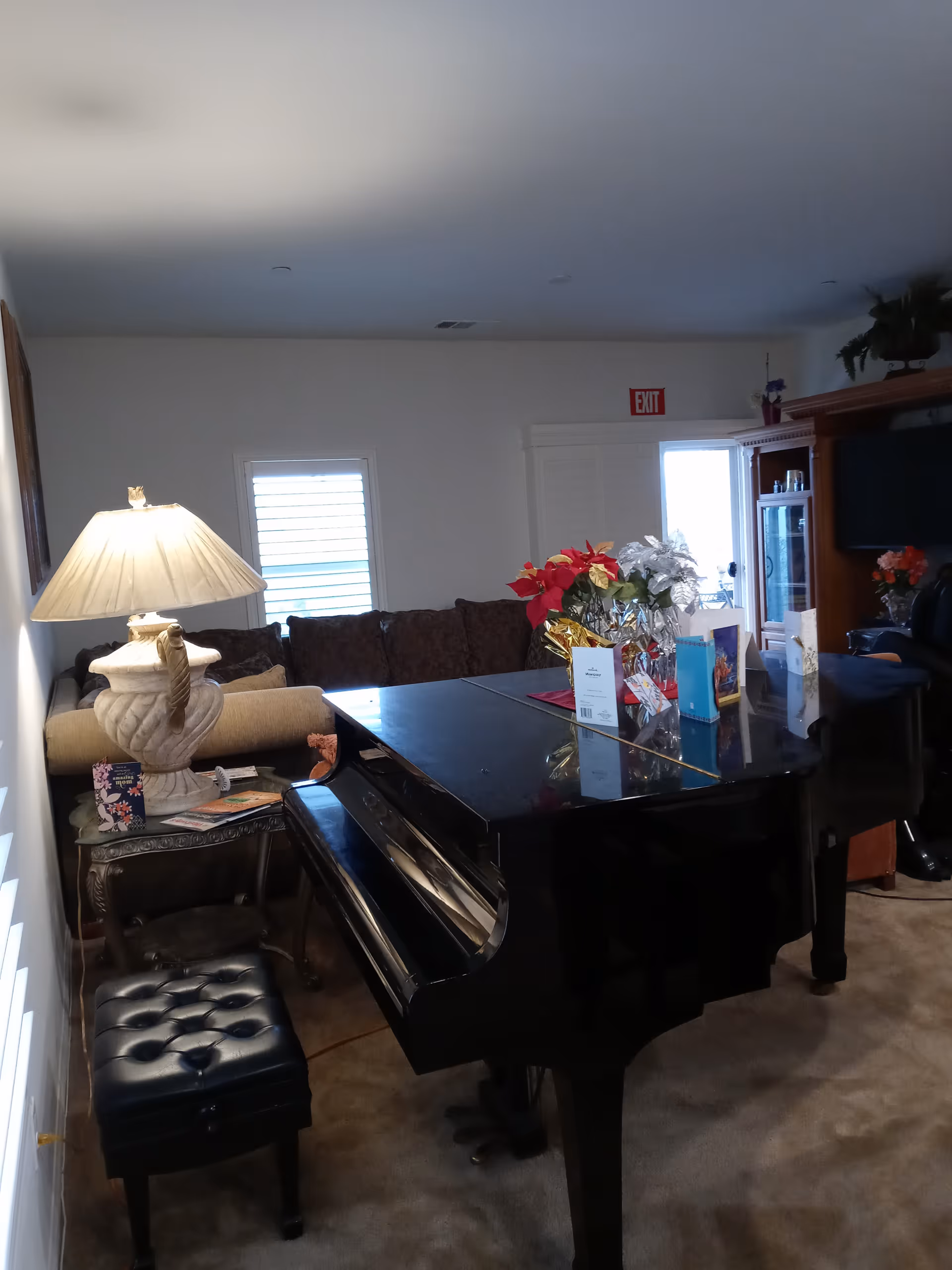Interior view of a living room with a black grand piano in the foreground, a cushioned piano bench, a side table with a decorative lamp, a brown sofa, and a wooden entertainment center with a TV. There are flowers and cards placed on the piano, and an exit sign is visible above a door in the background.