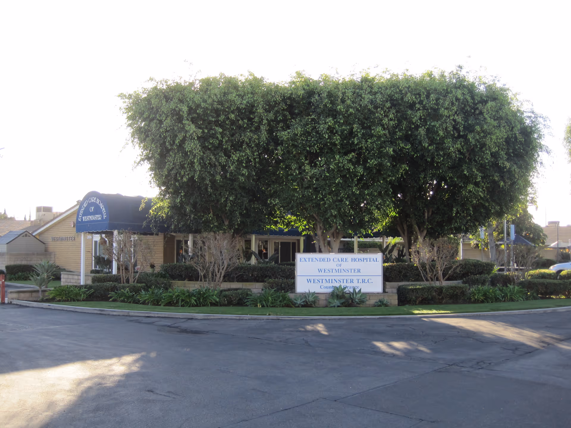 Front exterior of Extended Care Hospital of Westminster showing the entrance sign and large trimmed trees.