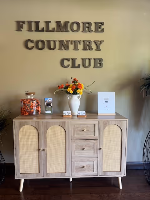 A wooden sideboard topped with a vase of flowers and decor beneath wall letters reading "FILLMORE COUNTRY CLUB".