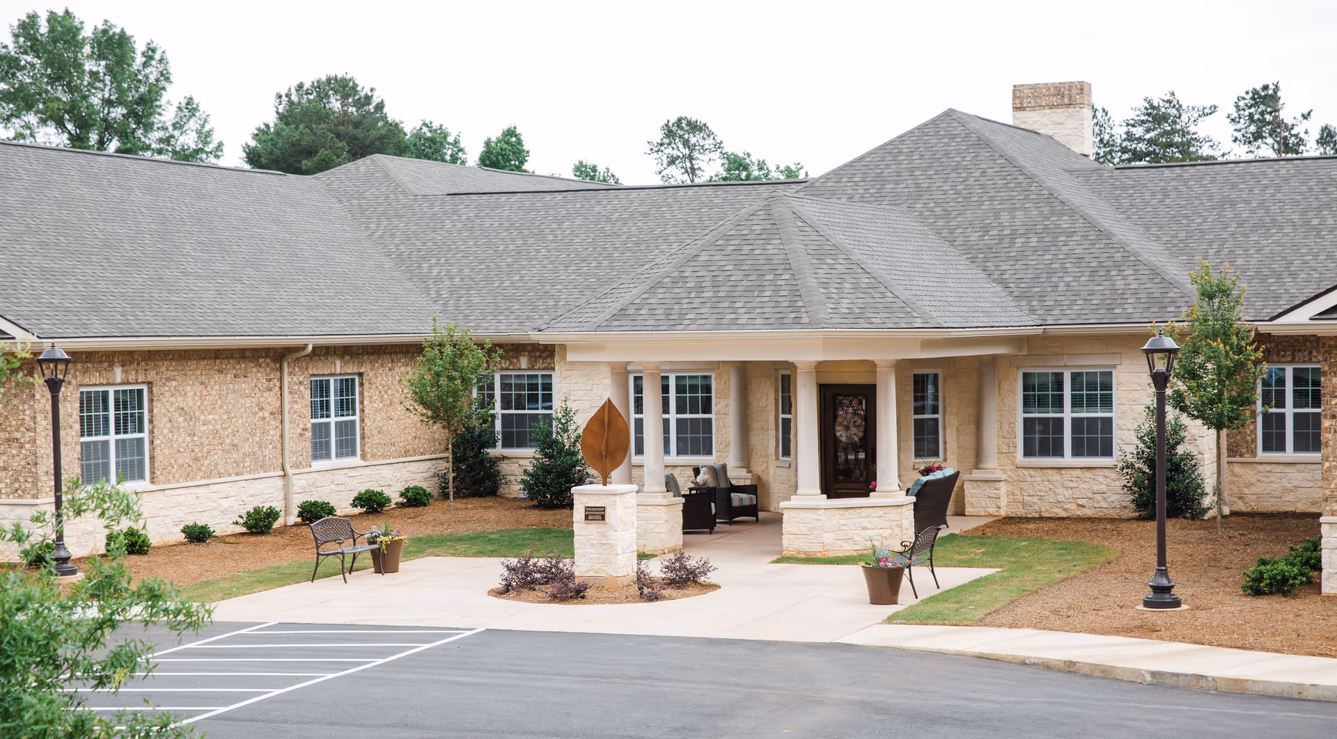 Exterior view of Lavender Hills Fort Mill facility showing a single-story building with a gray shingled roof, beige brick walls, several windows, a covered entrance with columns, outdoor seating, decorative plants, and a parking area in front.