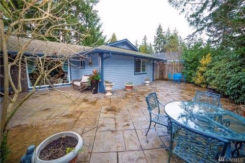 Outdoor patio area with stone tile flooring, a glass-top table with four metal chairs, potted plants, and a blue single-story building surrounded by trees and shrubs.