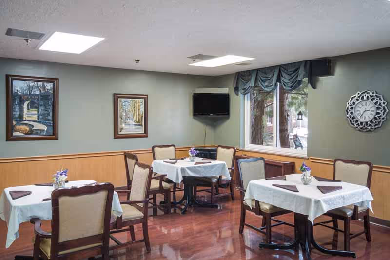 Small dining room with several tables set with white tablecloths and chairs, wall art, a TV, and a window.