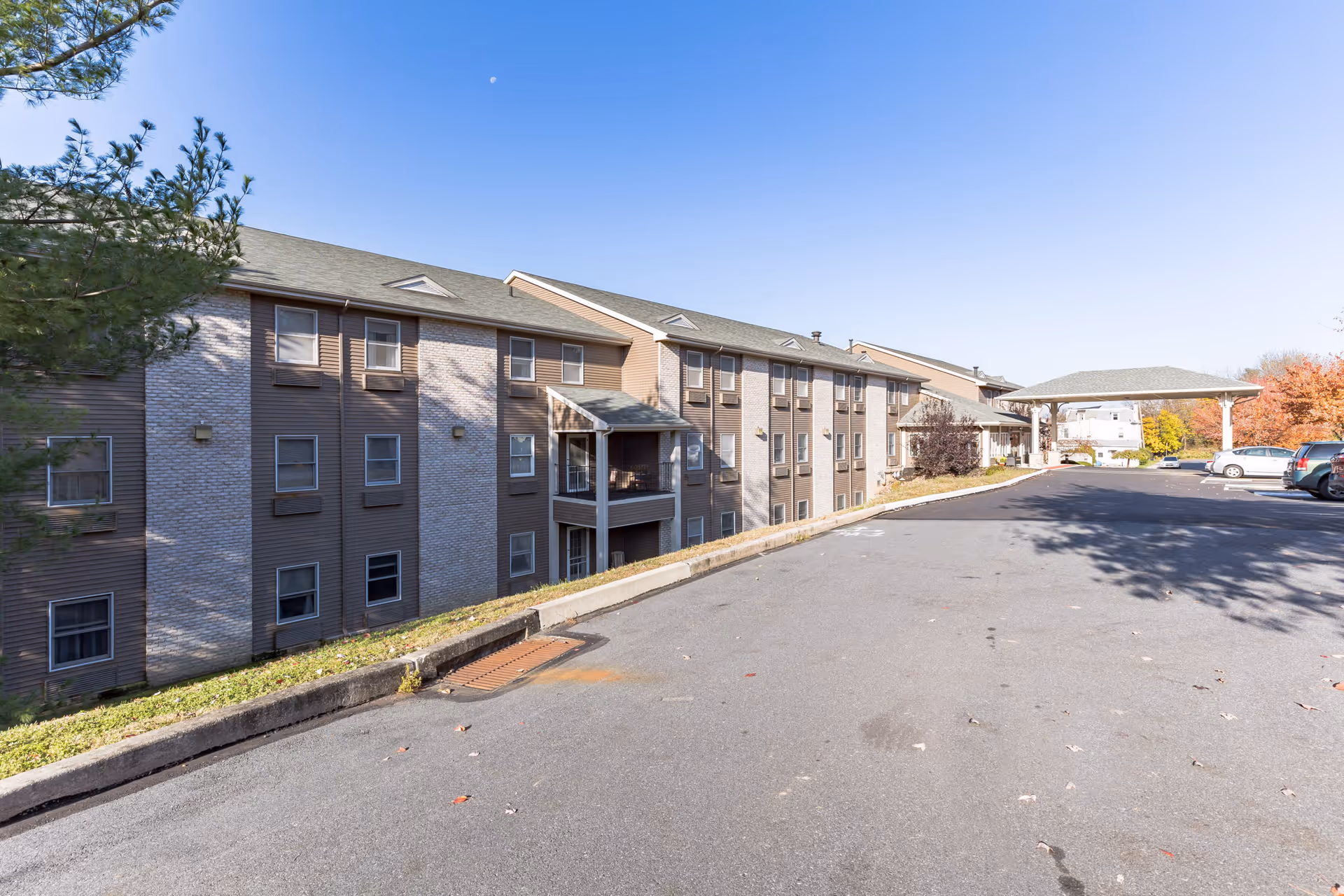 Exterior view of a three-story senior living building with a driveway, covered entry canopy, and parked cars under a clear sky.