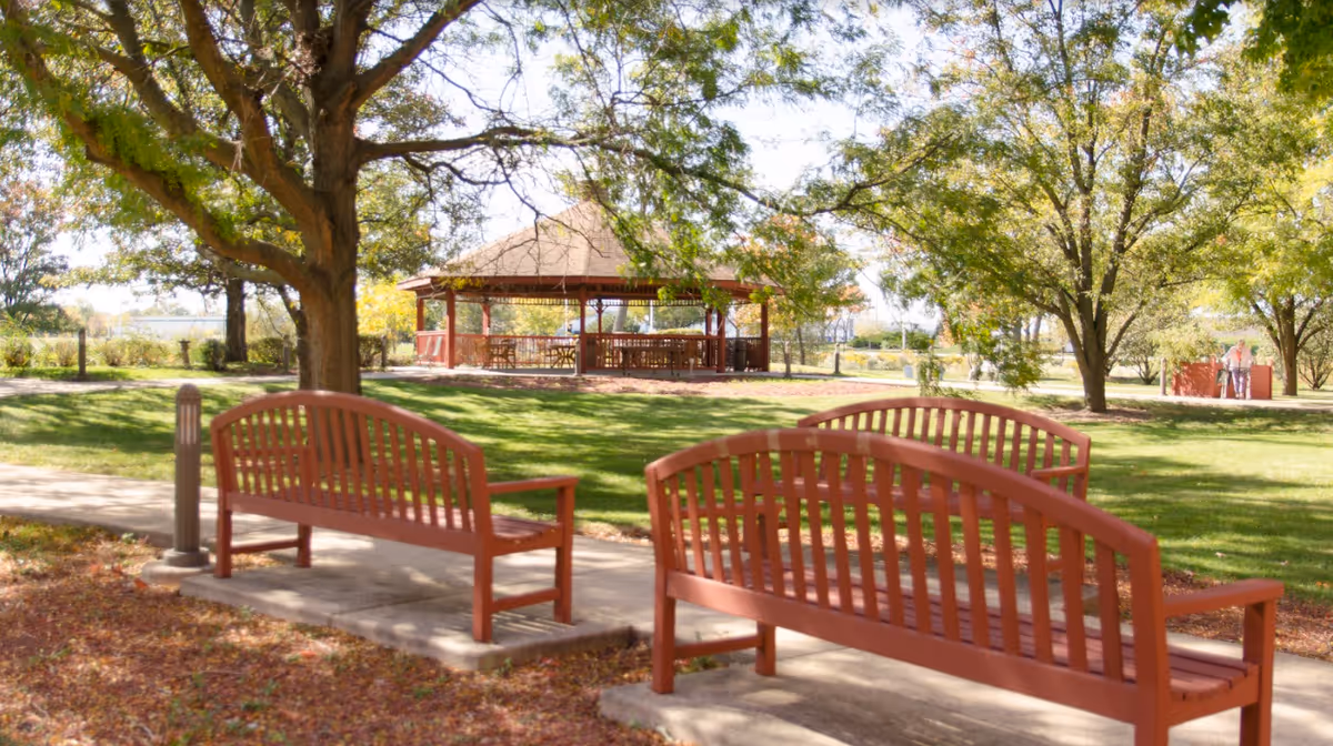 Outdoor park area with two wooden benches on a concrete path, large trees providing shade, and a gazebo with tables and chairs in the background. A person with a walker is visible in the distance near the trees.