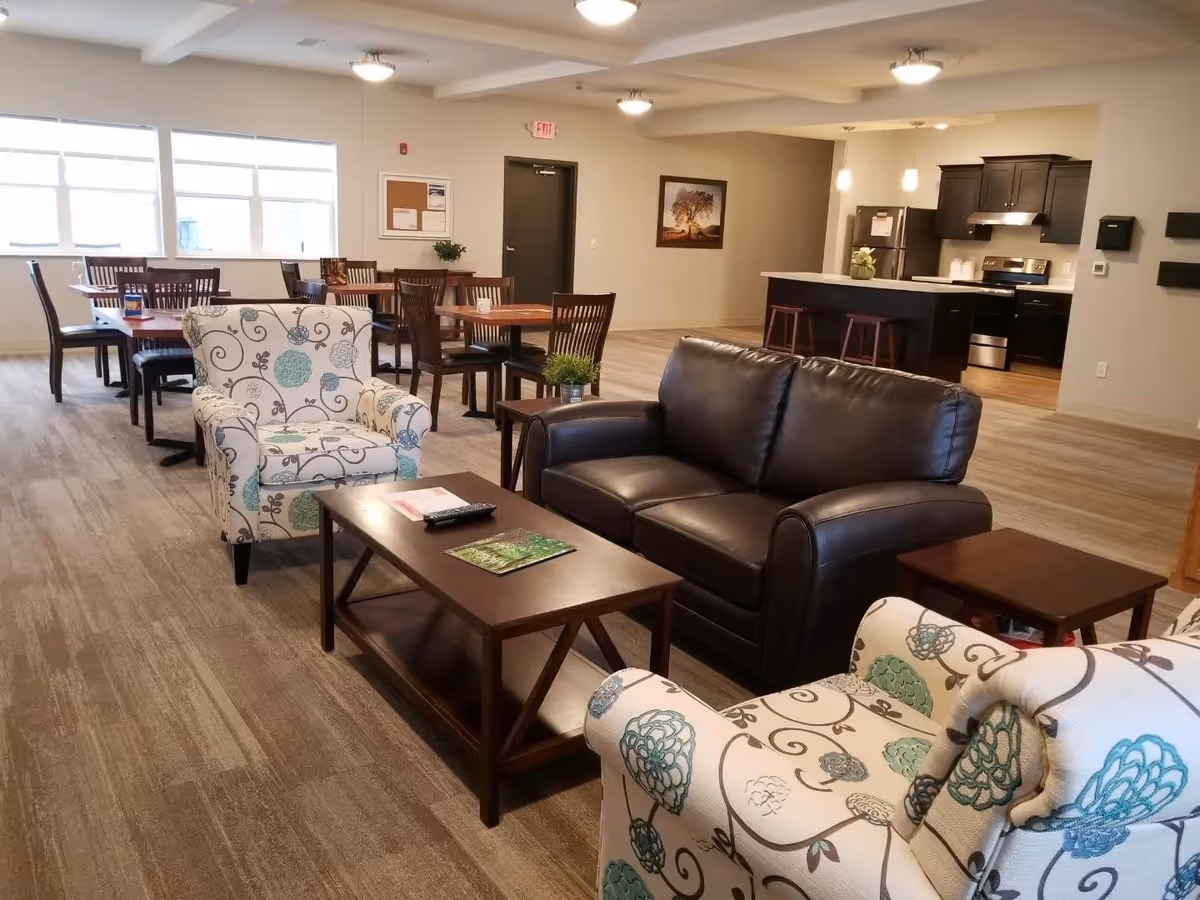 Open communal lounge with patterned armchairs, a leather sofa, coffee table, dining tables, and a kitchenette in the background.