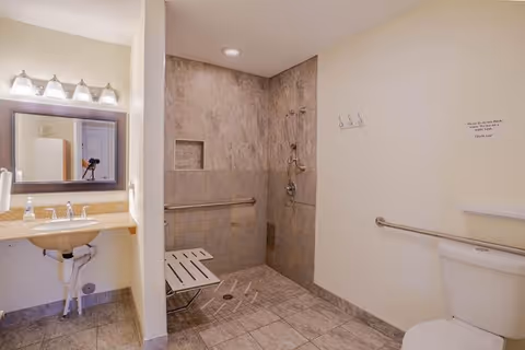 Accessible bathroom with a roll-in tiled shower, fold-down shower seat, grab bars, sink with mirror, and toilet.