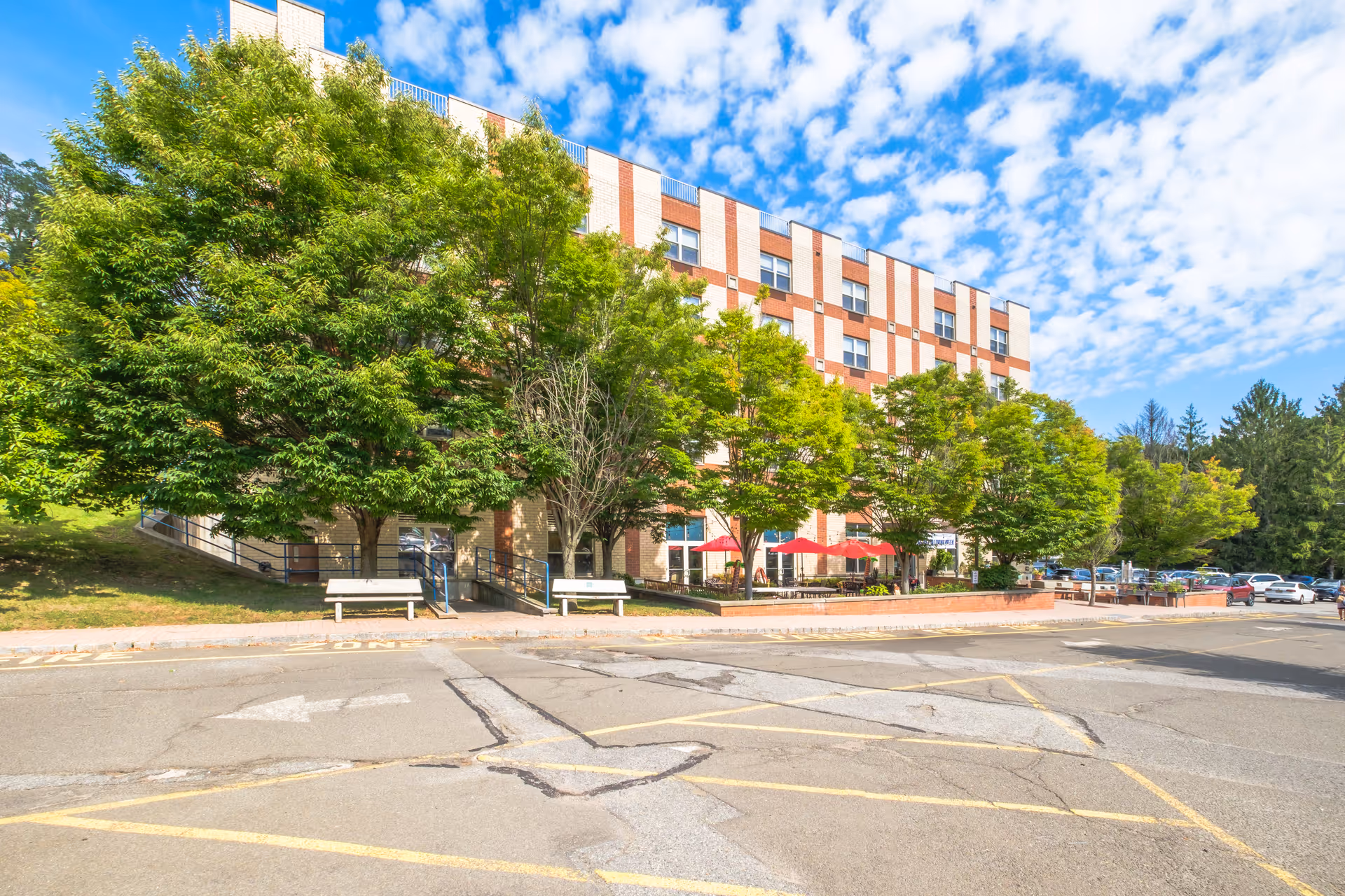 Exterior view of a multi-story brick building partially obscured by green trees with a parking lot in the foreground under a partly cloudy blue sky. There are benches and red umbrellas visible near the building entrance.