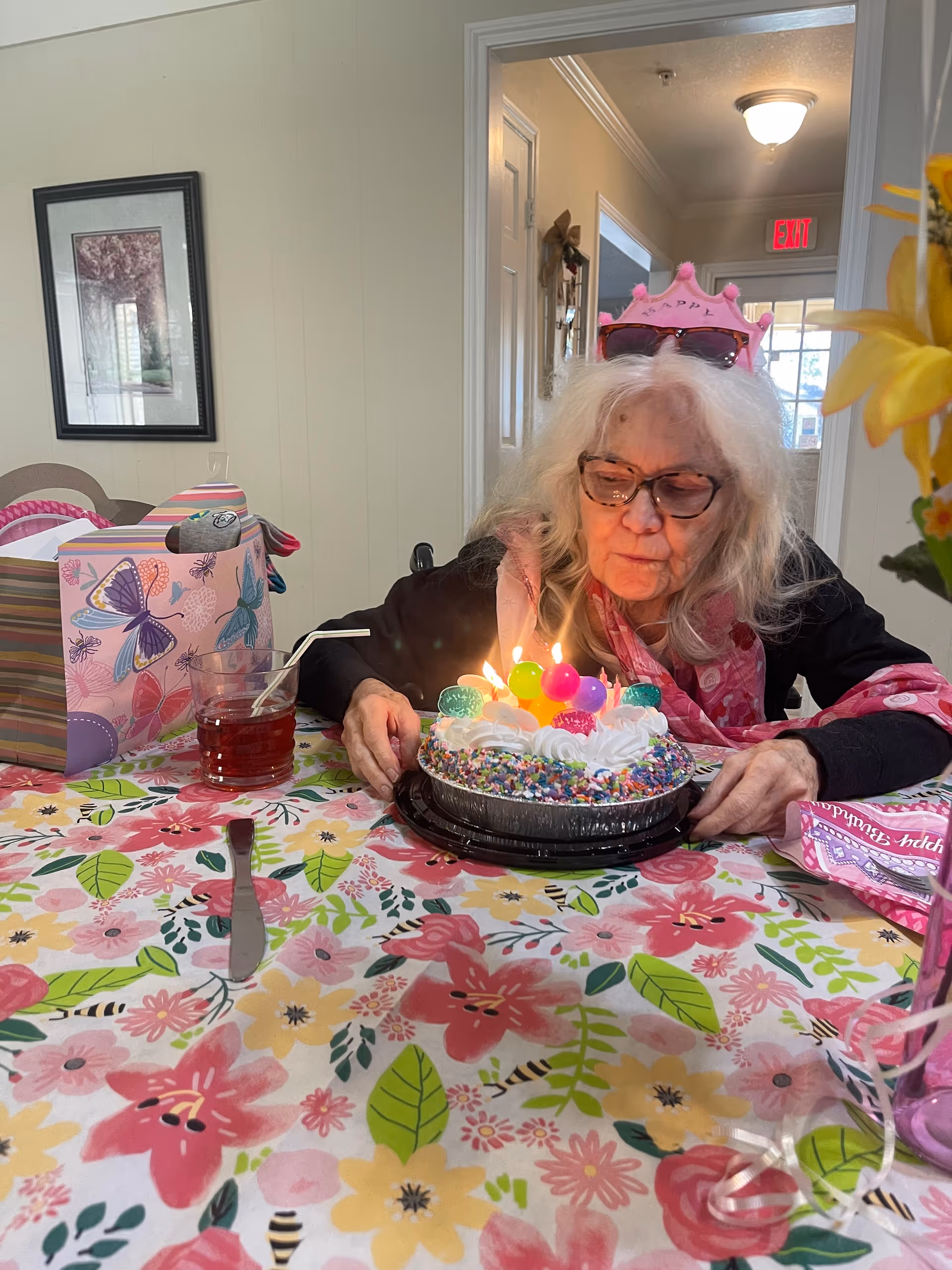 A woman wearing a pink paper crown leans over a small birthday cake with lit candles at a floral-covered table inside a communal room.
