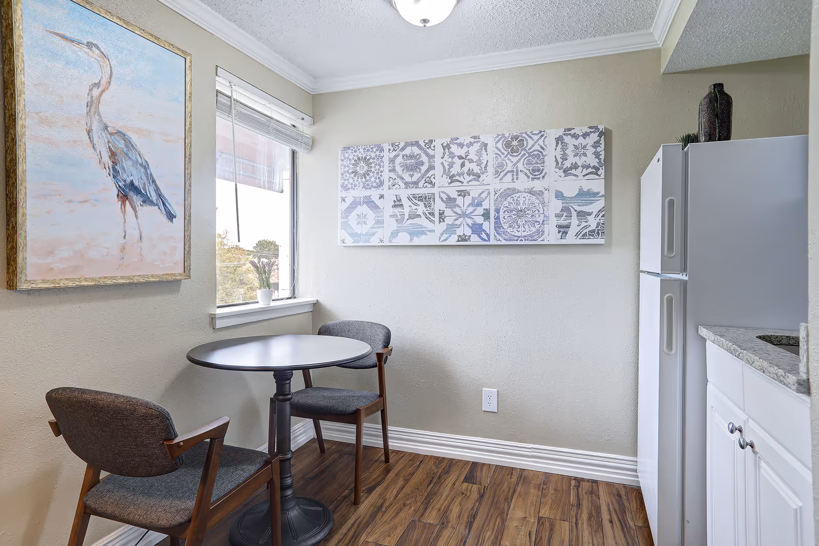 Small dining area with a round black table and two gray cushioned wooden chairs. There is a window with blinds partially open, a framed painting of a bird on the left wall, and a decorative tile-patterned artwork on the right wall. A white refrigerator and white cabinets with a granite countertop are visible on the right side. The floor is wood, and the walls are painted light beige.