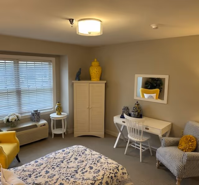 Bright bedroom with a patterned bed, white desk and chair, wardrobe, two armchairs, and yellow decorative accents.