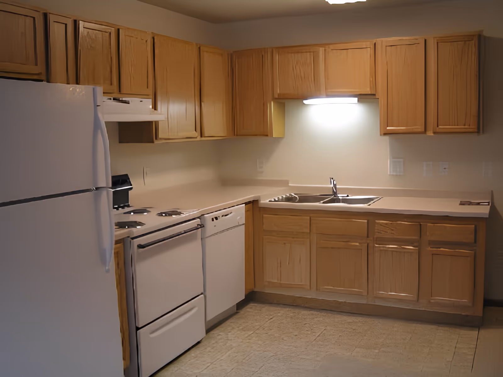 A kitchen with wooden cabinets, a white refrigerator, an electric stove with four burners, a dishwasher, and a double sink under a bright overhead light.