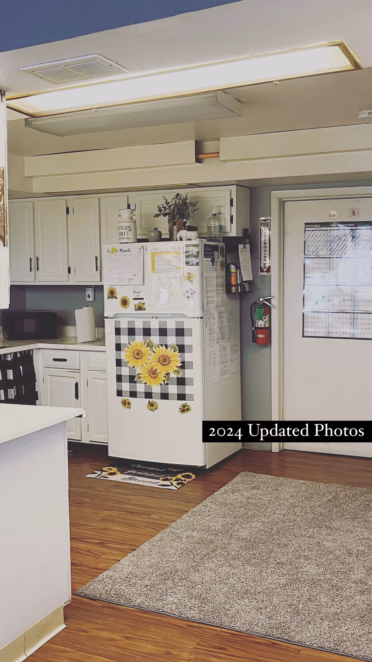 Interior view of a kitchen area with white cabinets, a refrigerator decorated with sunflower magnets and a black and white checkered cloth, a microwave on the counter, a fire extinguisher mounted on the wall next to a door, and a beige carpet on a wooden floor.