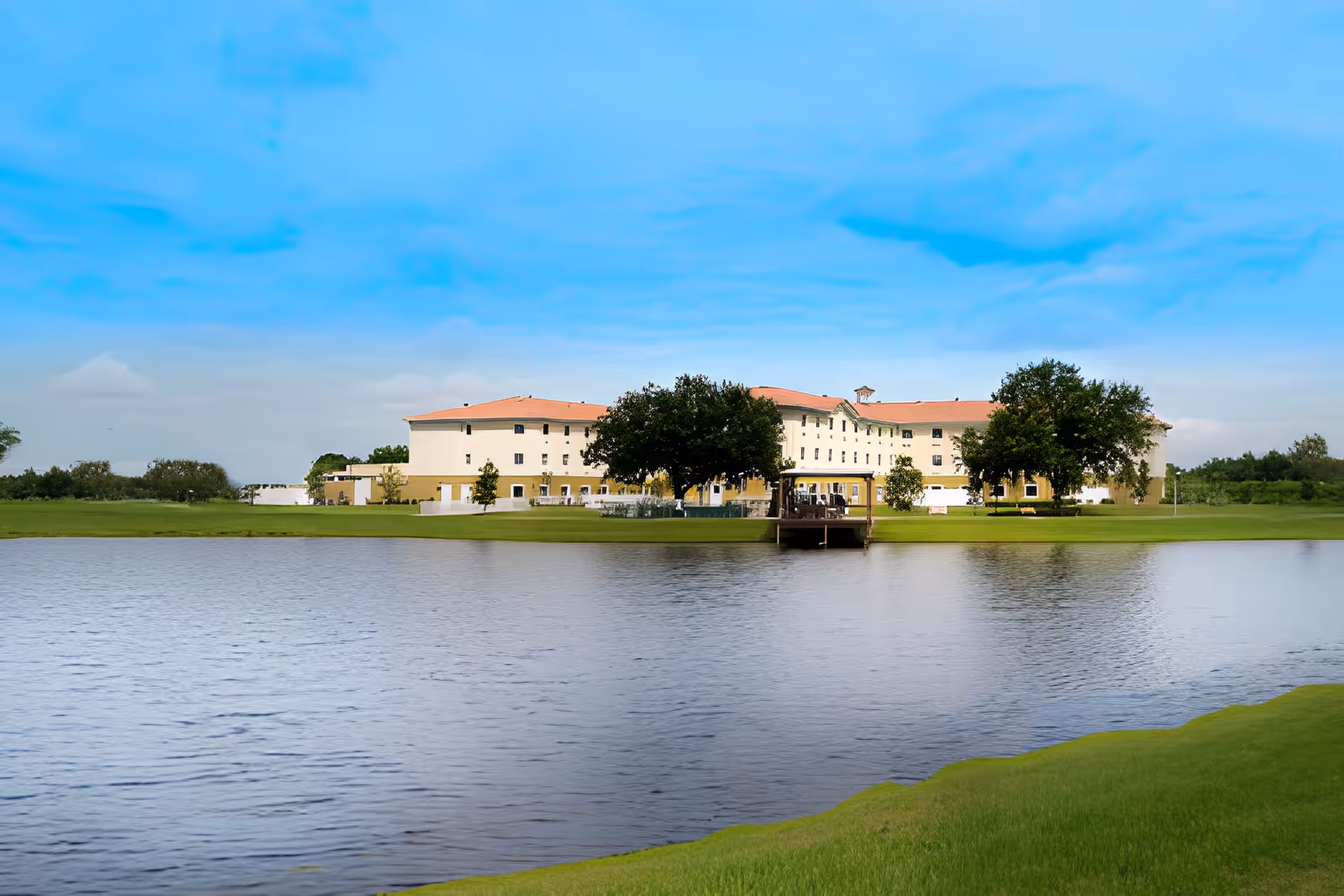 A large senior living facility building with a red-tiled roof situated behind a pond. The building is surrounded by green grass and trees under a blue sky with some clouds.