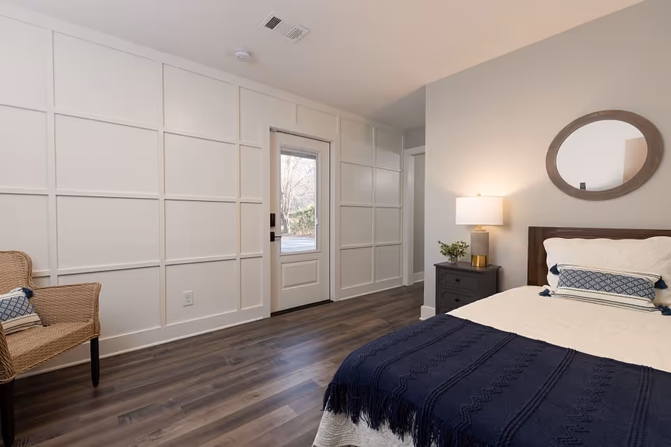 A bright bedroom with a bed covered in white and navy blue bedding, a dark wooden nightstand with a lamp and small plant, a round mirror on the wall above the bed, a wicker chair with a cushion, and a door with a window leading outside. The room has white paneled walls and dark wood flooring.