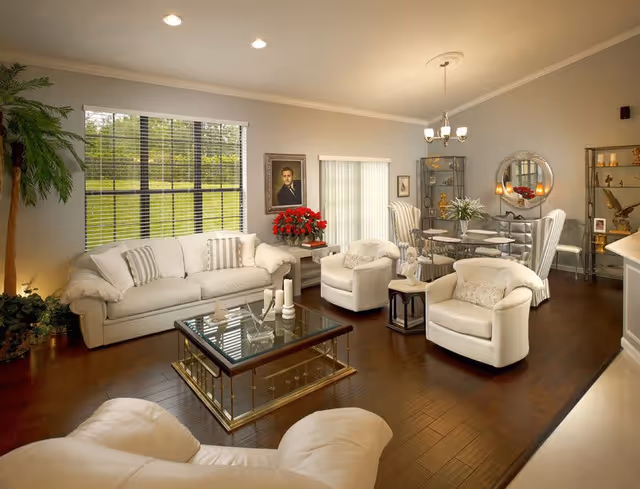 Bright, elegantly furnished living and dining area with white sofas and armchairs, a glass coffee table, hardwood floors and a large window overlooking a green lawn.