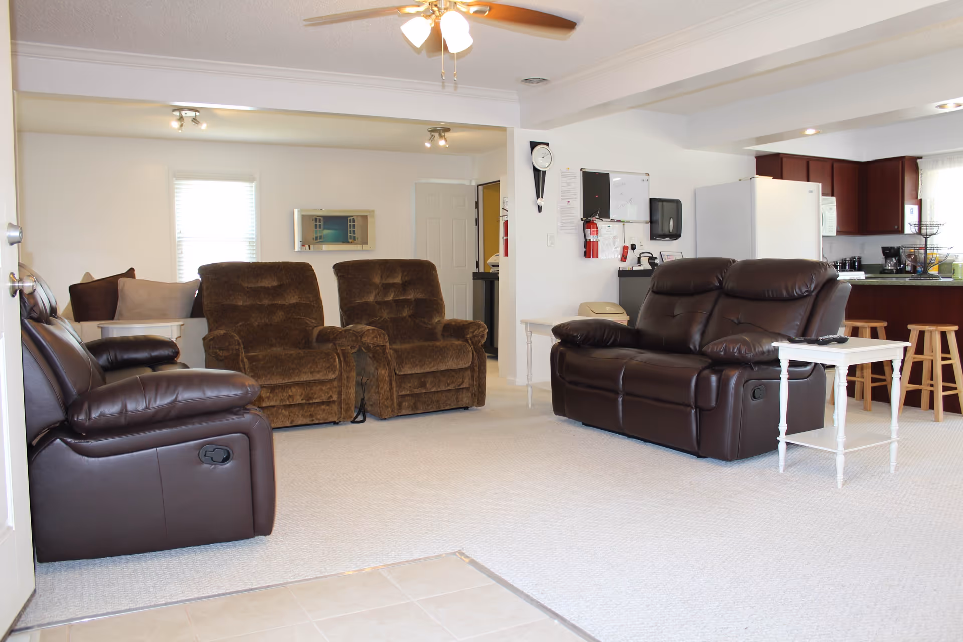 A spacious living room area with two brown recliner chairs, a dark brown leather loveseat, and a matching recliner chair. There are side tables next to the seating, a ceiling fan with lights, and a kitchen area with wooden stools and white appliances in the background.