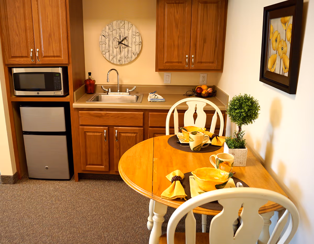 A small kitchen and dining area with wooden cabinets, a stainless steel microwave and mini fridge, a sink, and a round wooden table set for two with yellow napkins, bowls, mugs, and a small potted plant. A decorative wall clock and framed yellow flower artwork hang on the walls.