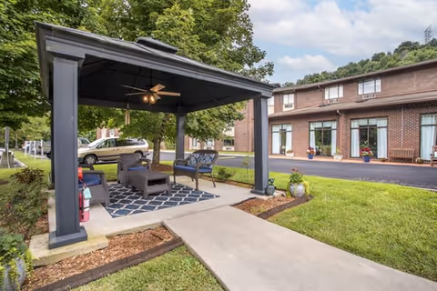 Outdoor seating area under a black gazebo with a ceiling fan, cushioned chairs, a small table, and a patterned rug. The gazebo is situated on a concrete path surrounded by grass and landscaping. In the background, there is a two-story brick building with large windows and a paved driveway.