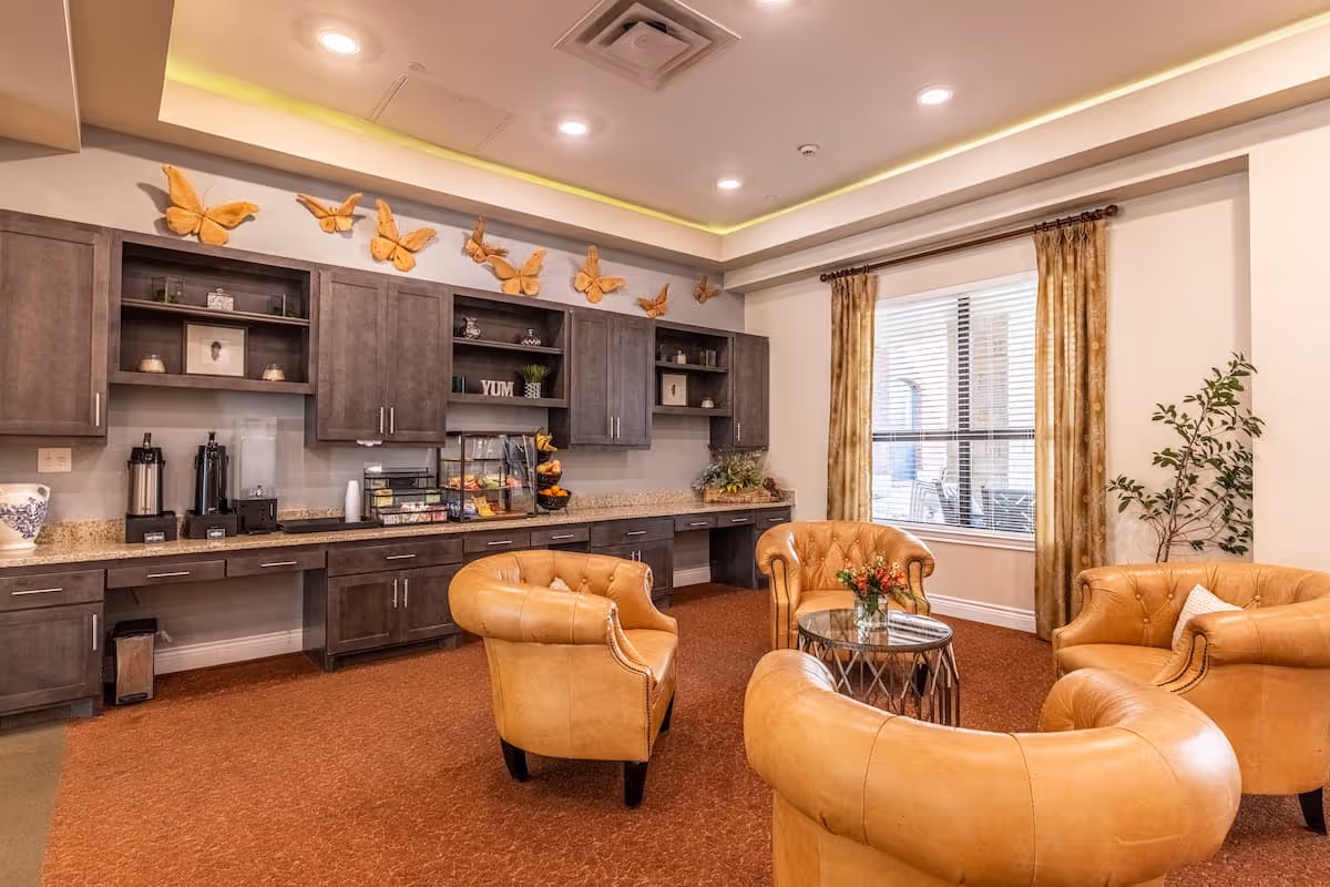 A cozy common area in an assisted living facility featuring four tan leather armchairs arranged around a small glass-top table with a flower arrangement. Behind the seating area is a long counter with dark wooden cabinets above and below, decorated with orange butterfly wall art and various small decor items. A large window with gold curtains lets in natural light, and a potted plant is placed in the corner.