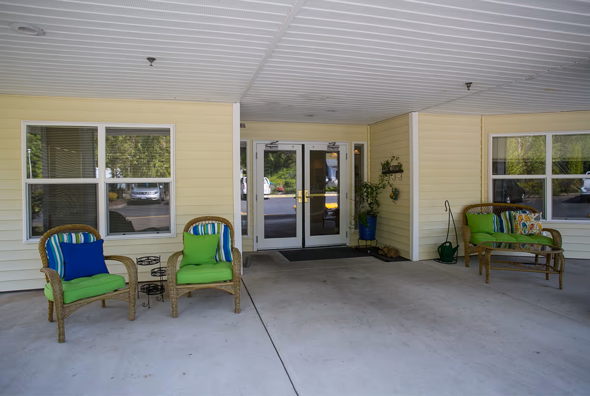 Covered entrance area of a senior living facility with two wicker chairs with green and blue cushions on the left, a wicker loveseat with green and patterned cushions on the right, a glass-top wicker coffee table, and double glass doors in the center. There is a welcome sign on the wall next to a potted plant.