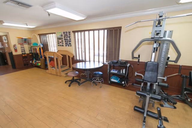 A rehabilitation room in a senior living facility with exercise equipment including a multi-gym machine on the right, a small round table with two stools in the center, and various therapy tools and equipment along the left wall. The room has wooden flooring, large windows with vertical blinds, and beige walls with wood paneling on the lower half.