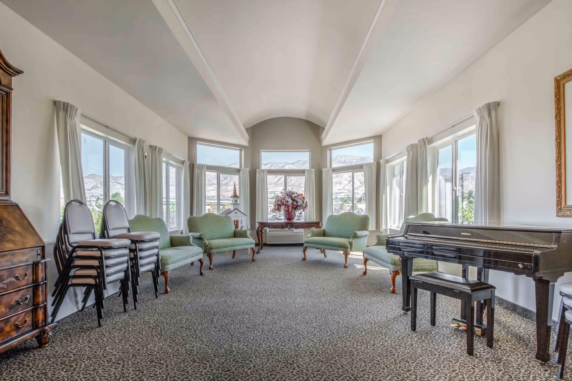 A bright and spacious living room with large windows offering a view of mountains. The room features several green upholstered armchairs arranged along the walls, a black grand piano with a matching bench on the right side, a wooden cabinet on the left, and a table with a large floral arrangement in the center near the windows. Stacks of chairs are placed against the left wall.