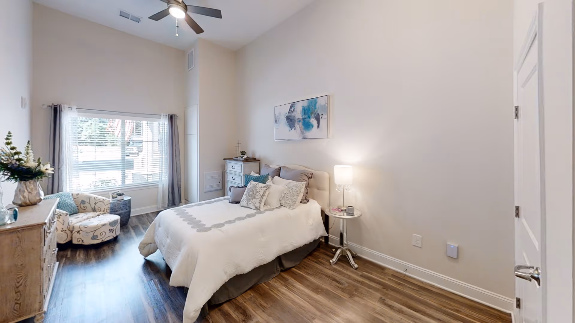 A bright and airy bedroom with a large window covered by sheer curtains, a bed with white and gray bedding and multiple pillows, a small round side table with a lamp, a wooden dresser with a vase of flowers, and a patterned armchair next to a small round table. The room has wood flooring and a ceiling fan with a light.