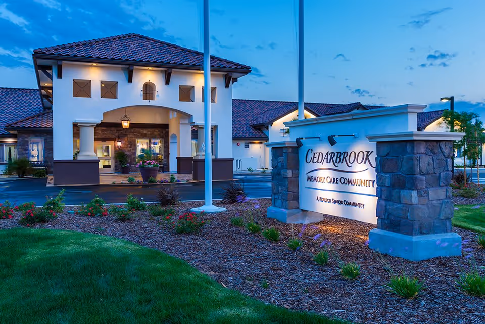 Exterior view of Cedarbrook Memory Care Community building at dusk with well-lit entrance, landscaped garden, and a prominent stone sign displaying the facility's name.